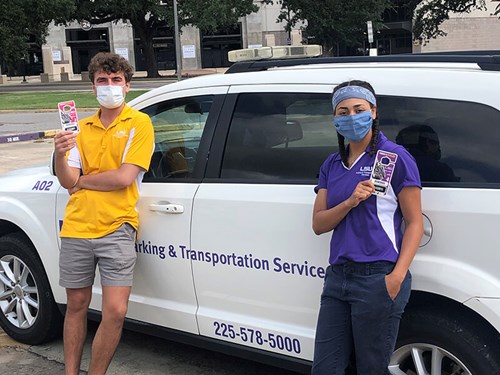 A man in a yellow shirt and a woman in a purple shirt wear face masks and hold parking permits whilst standing in front of a white car.